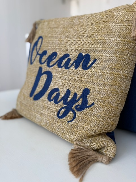 A jute cushion embroidered with 'Ocean Days' in navy script, with tassels at the corners.